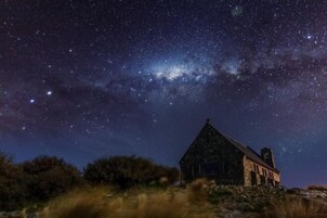 Edificio de piedra rústico con techo puntiagudo, rodeado de hierba y arbustos, bajo un cielo estrellado.