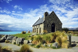 A stone church with a steep roof, situated on a grassy area with rocks and shrubs, overlooking a body of water.
