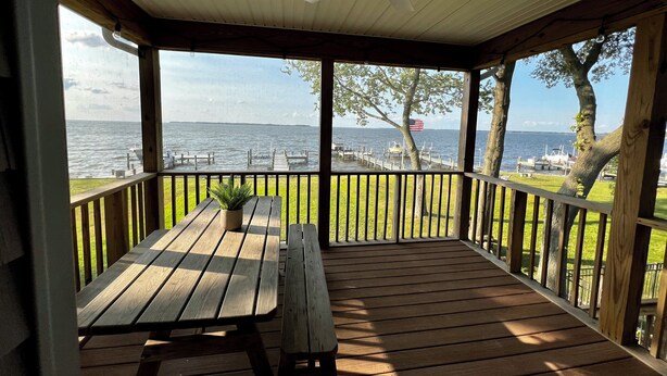 A covered outdoor deck with a wooden table and bench, overlooking a body of water with docks and boats.