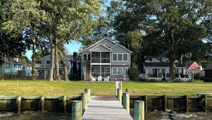 A wooden pier leading to a house with a balcony, surrounded by trees and a grassy area.