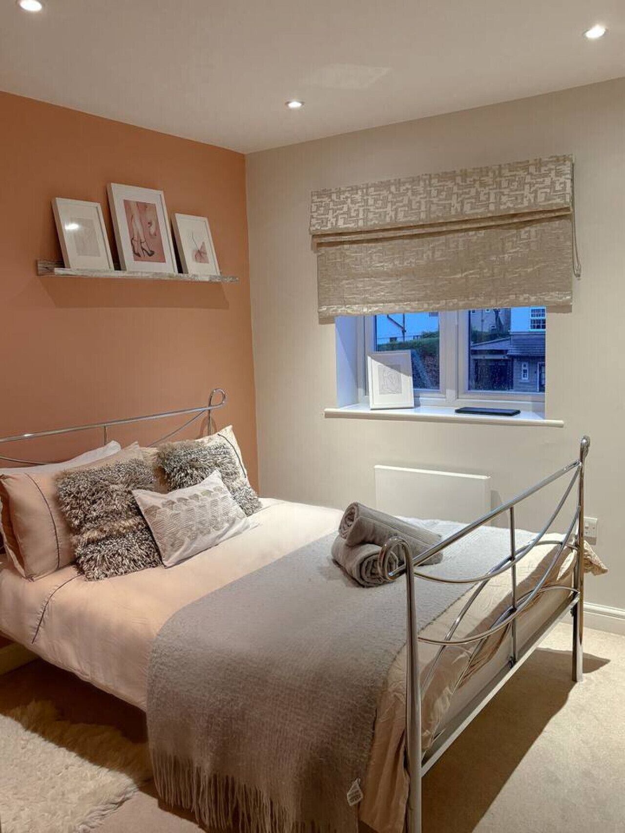 Un dormitorio con estructura de cama de metal, una ventana con persiana color beige y un estante de pared con cuadros enmarcados.
