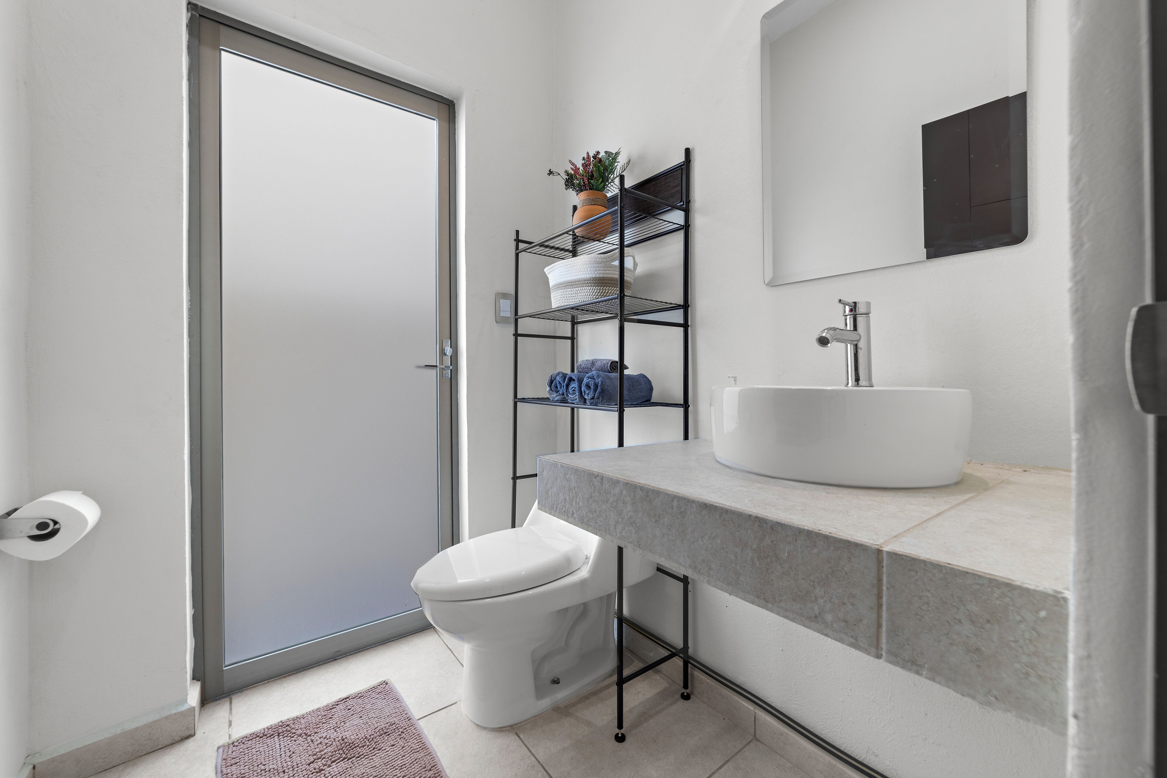 Un baño moderno con inodoro, lavabo, espejo y estante con una planta.