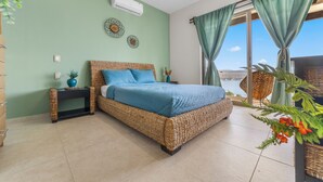 A bedroom with a wicker bed, a nightstand, a potted plant, and a view of the sea through a window with curtains. - Casa Familiar Alberca vista al lago 4 RC (Jojutla)