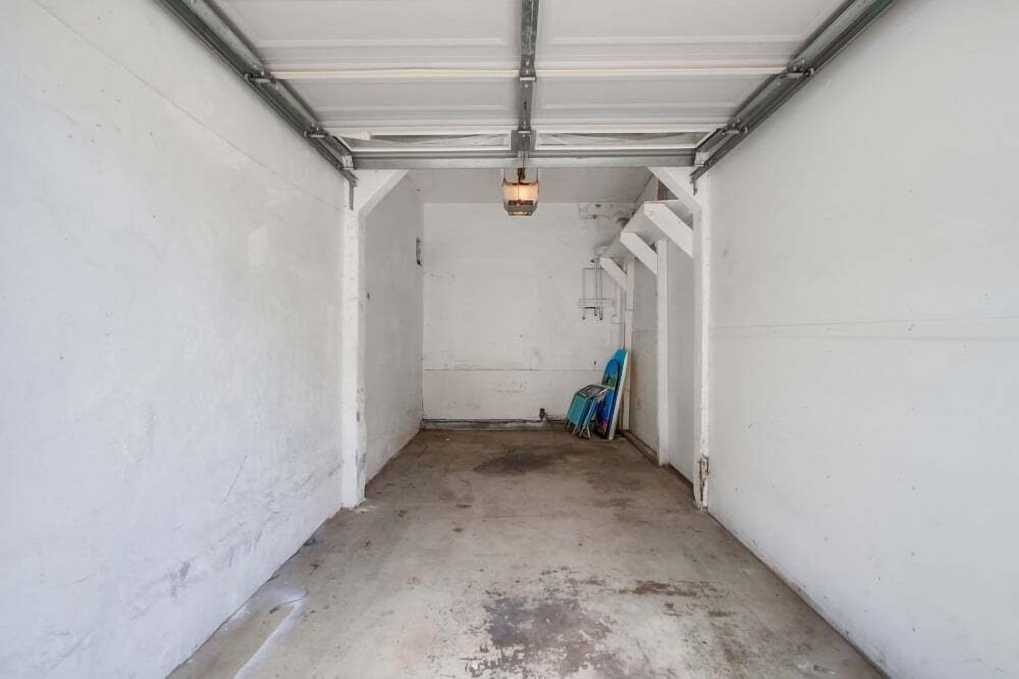 A garage with a concrete floor and white walls, featuring a single light fixture and a blue object leaning against the wall.
