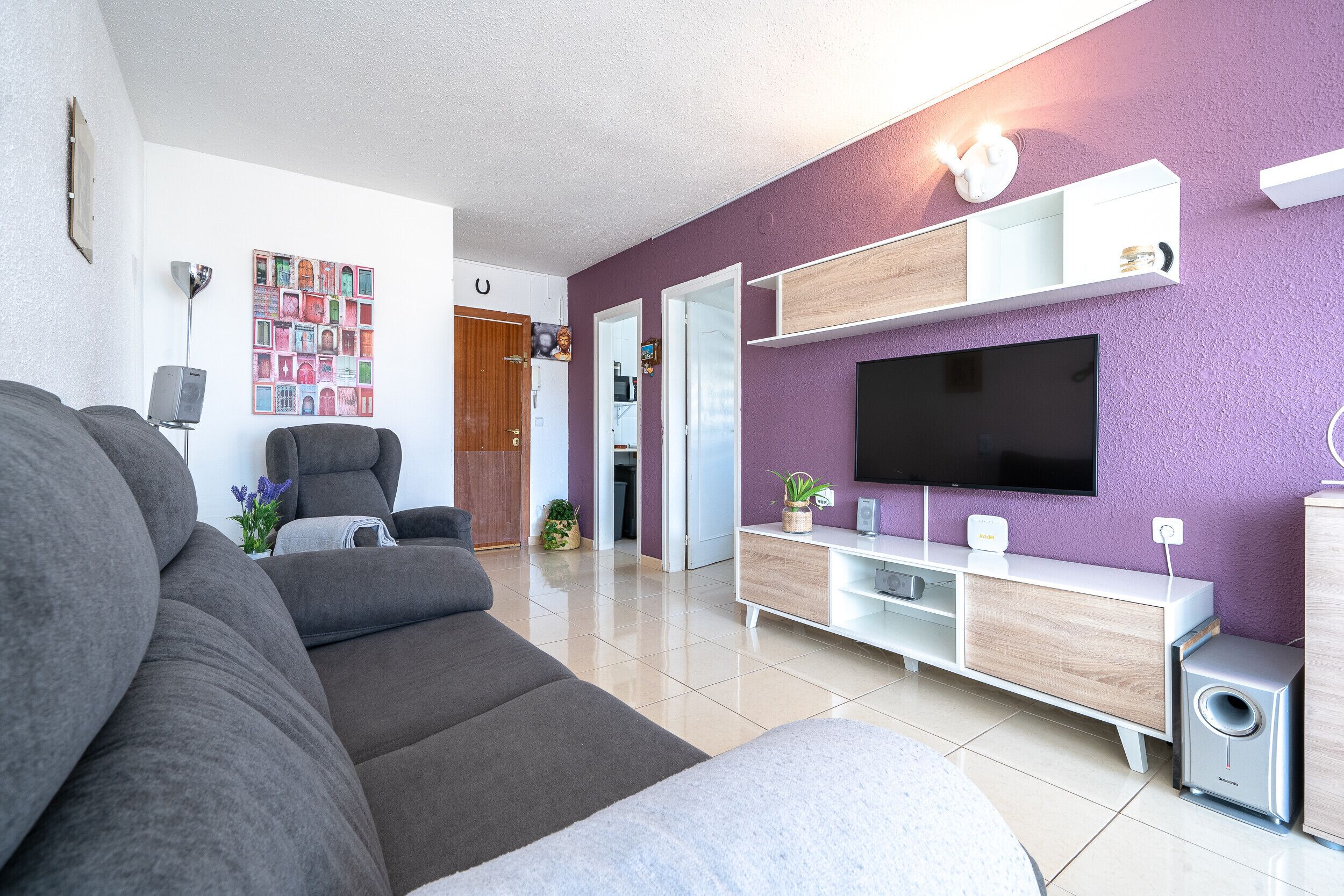 A modern living room with a large grey sofa, a flat-screen TV mounted on a purple wall, and a white entertainment unit with a speaker.