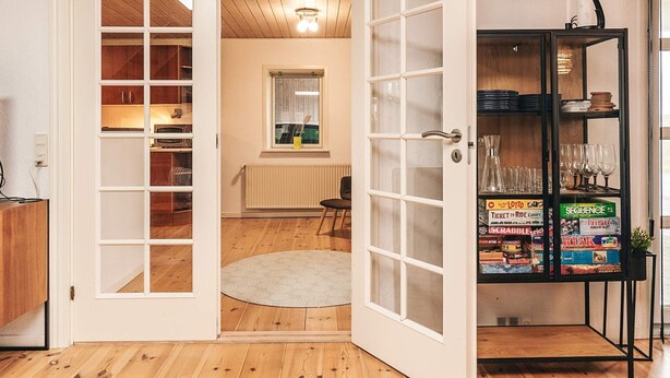 A room with a wooden floor, a white door with glass panels, a radiator, a small window, and a shelf with board games. - 6 Person Holiday Home in Snedsted (Snedsted)