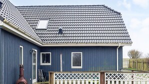 A blue house with a tiled roof, a chimney, and a wooden deck with a white fence. - 6 Person Holiday Home in Snedsted (Snedsted)