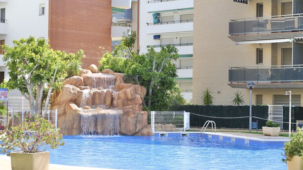 A swimming pool with a waterfall feature, surrounded by greenery and a fence. - MALDIVAS III Complex (La Pineda)