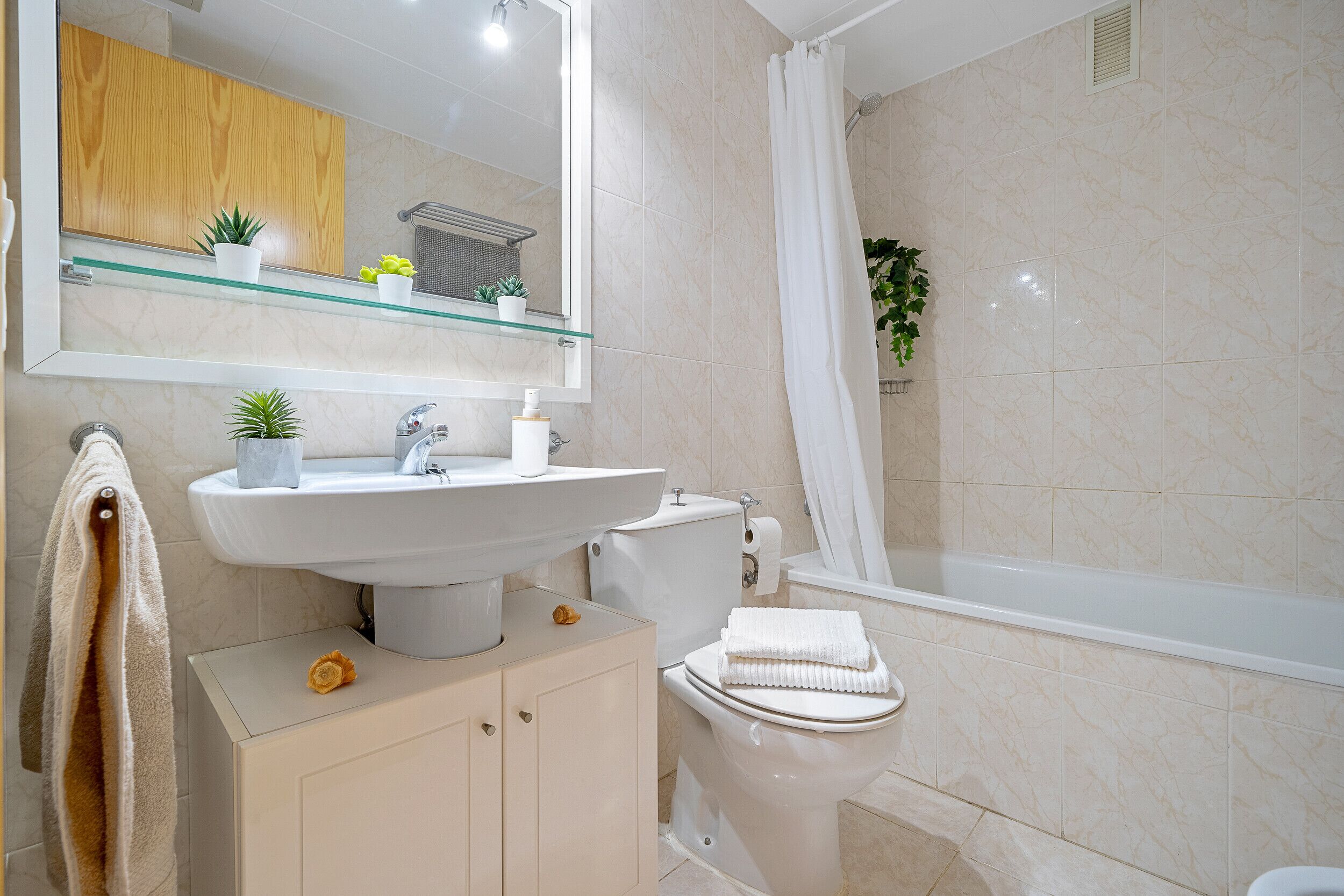 Un baño con lavamanos, inodoro y bañera. Hay plantas en macetas en el estante sobre el lavamanos.