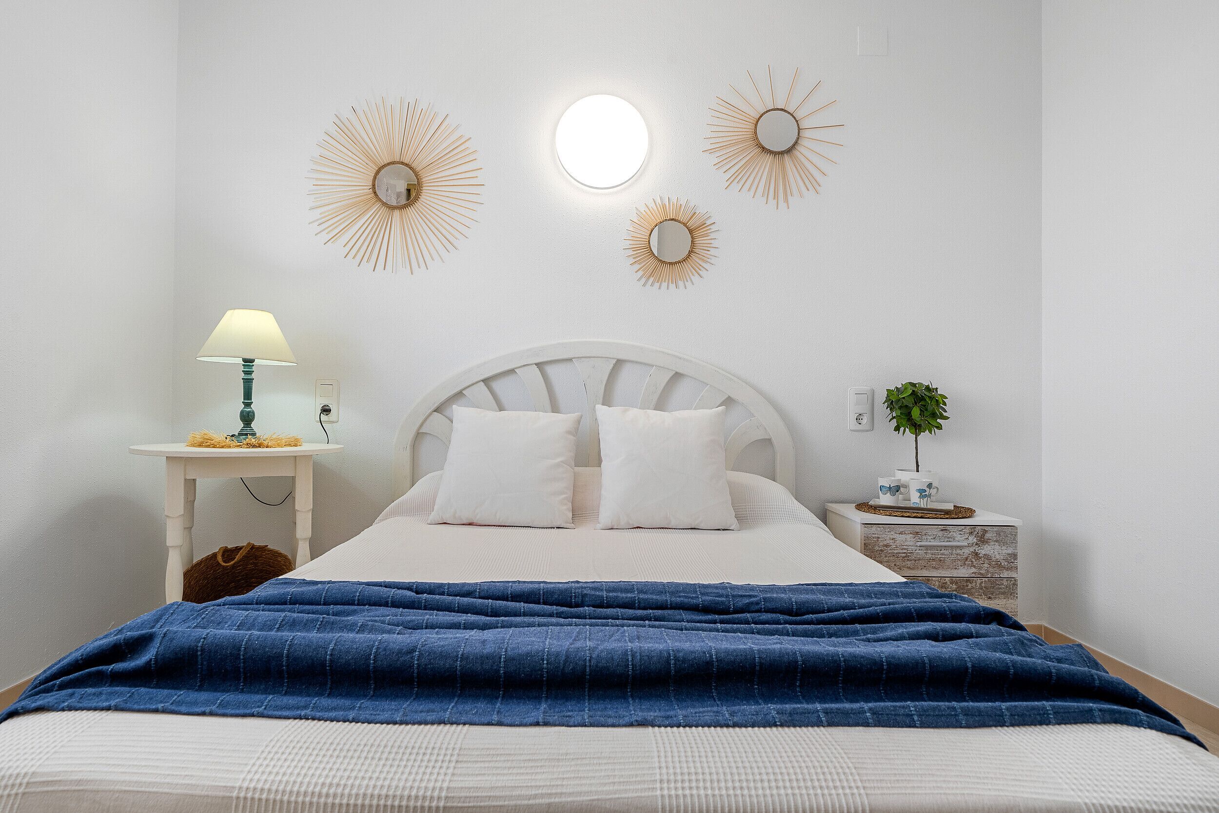 A bedroom with a bed, a nightstand with a lamp, a wall-mounted mirror, and decorative wall sconces.