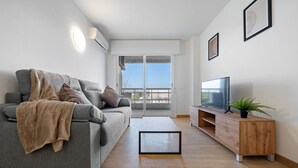 A modern living room with a grey sofa, a wooden TV stand, and a balcony with a glass door. - AQUAMARINA II Apartments (La Pineda)
