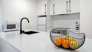 A modern kitchen with white cabinetry, a black sink, a microwave, and a fruit basket on the counter. - AQUAMARINA II Apartments (La Pineda)