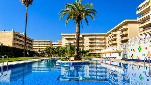 A swimming pool with tiled flooring, surrounded by palm trees and a multi-story building with balconies. - AQUAMARINA II Apartments (La Pineda)