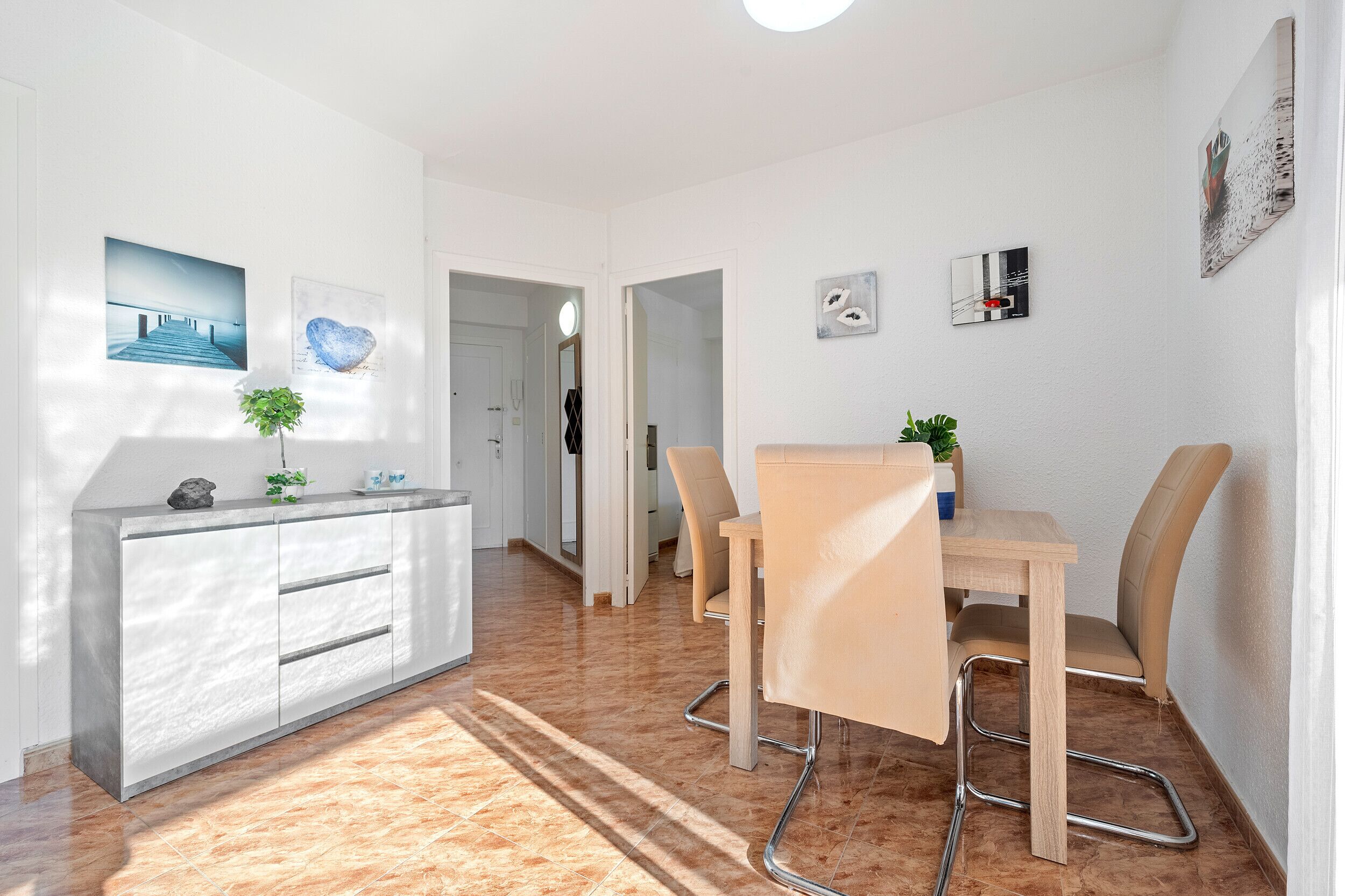 Une chambre d’hôtel moderne avec un coin repas, une console blanche dotée de tiroirs, une petite plante en pot et une vue sur le couloir.