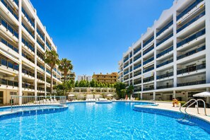 A swimming pool surrounded by multi-story buildings with balconies, palm trees, and a clear blue sky. - ROYAL APARTMENTS SALOU (Salou)