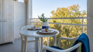Ein Balkon mit einem weißen Tisch für zwei Personen, einem Stuhl mit blauem Handtuch und Blick auf Bäume und Gebäude.