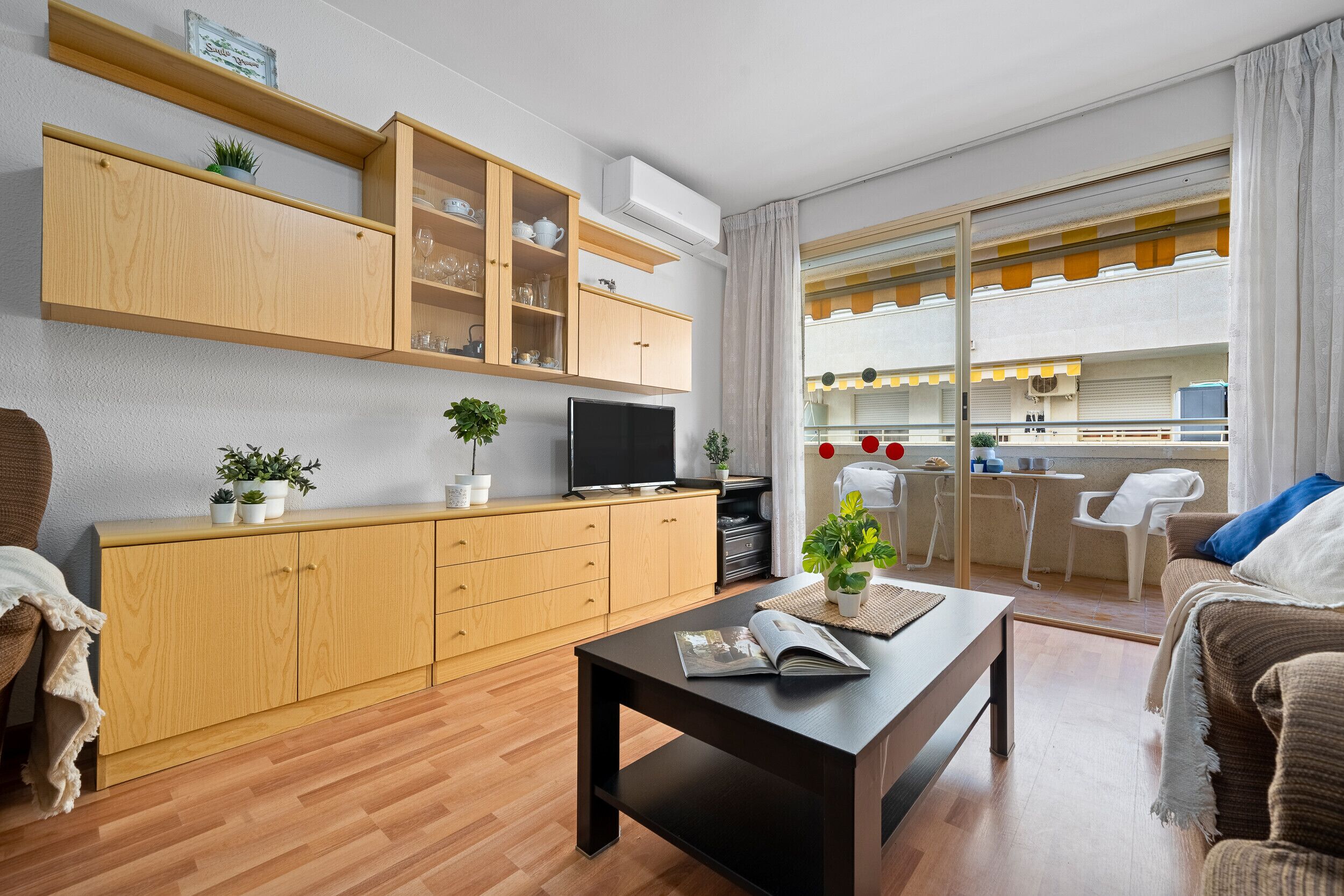 Un salón moderno con armarios de madera, un televisor de pantalla plana, una mesa de centro con una revista y vista a la cocina a través de una puerta de cristal.