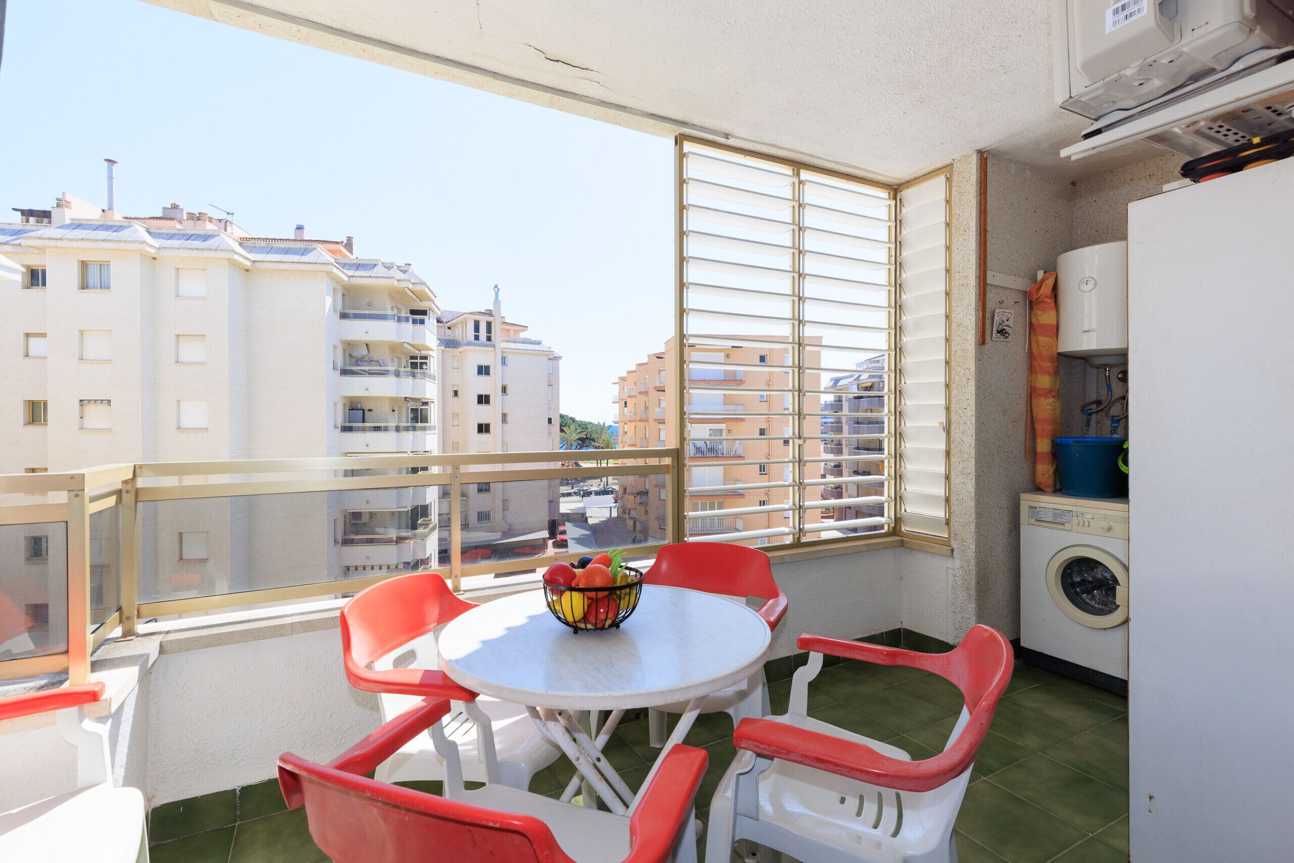 Un balcón con mesa y sillas, lavadora y ventana con persianas.