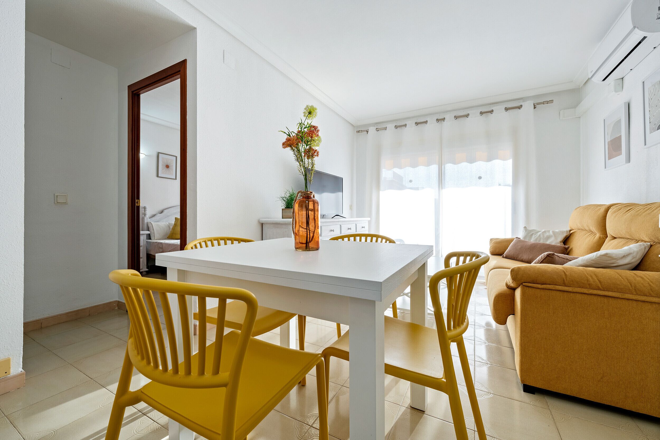 A living room with a white table and yellow chairs, a sofa, and a vase with flowers.