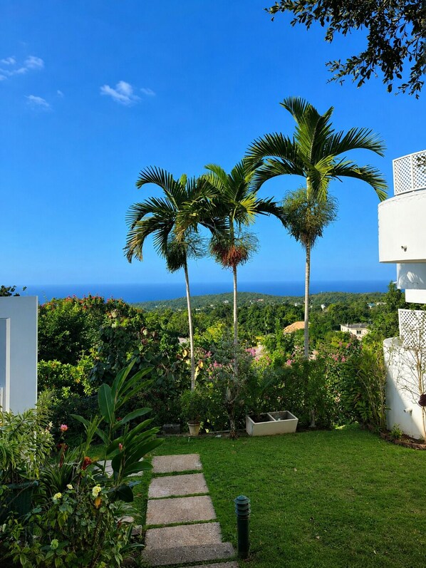 A garden with a stone pathway leading to a white building, surrounded by lush greenery and palm trees, with a view of the ocean in the background.