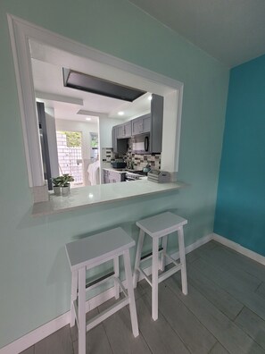 A modern kitchen with a central island, white bar stools, and a view into the adjacent living area.