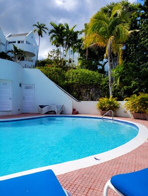 Piscina com água azul cristalina, rodeada por um edifício branco, palmeiras e um caminho de tijolos vermelhos.
