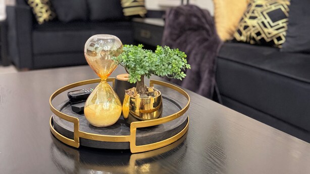 Un salon moderne avec un canapé noir, des coussins décoratifs, une petite plante en pot, ainsi qu’un ensemble original composé d’un sablier et d’un vase, le tout posé sur un plateau noir et doré.
