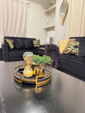 Una sala moderna con un sofá negro, cojines decorativos, una pequeña planta en maceta y un original juego de ampulleta y jarra sobre una bandeja negra y dorada.