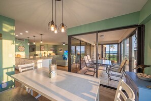 Een moderne keuken met een centraal eiland, een eethoek met tafel en stoelen, en uitzicht op de oceaan door grote schuifdeuren.