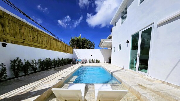 A modern outdoor swimming pool with two white sun loungers, surrounded by a white wall and a wooden fence.