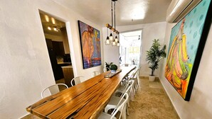 A long wooden dining table with white chairs, surrounded by two large paintings, in a well-lit room with a tiled floor.