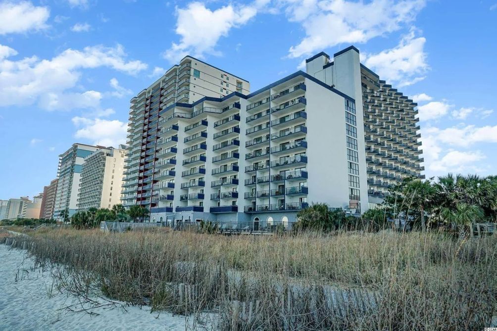 A high-rise hotel with multiple balconies, located near a beach with sand and sparse vegetation.
