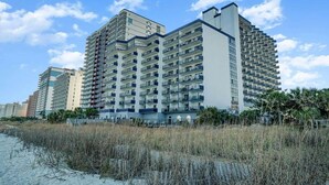 A high-rise hotel with multiple balconies, located near a beach with sand and sparse vegetation.
