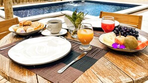 Uma mesa de madeira com itens de café da manhã, um copo de suco de laranja e vista para uma piscina e praia.