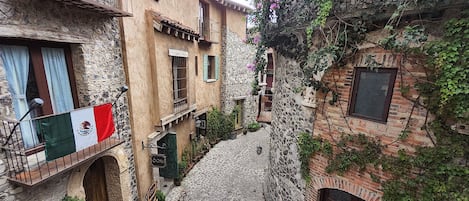 A narrow cobblestone street flanked by stone buildings with balconies, a flag, and potted plants.
