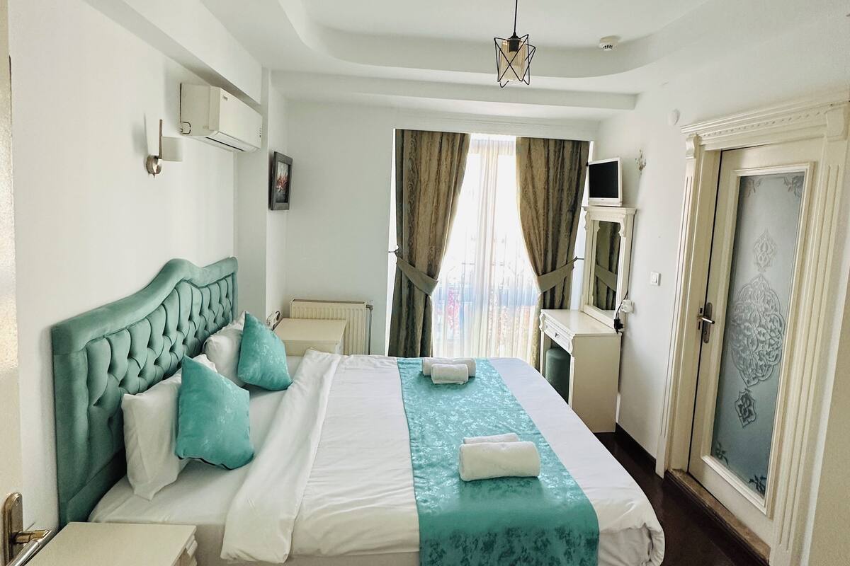 A hotel room with a large bed, a desk, a wardrobe, and a view of the city through the window.