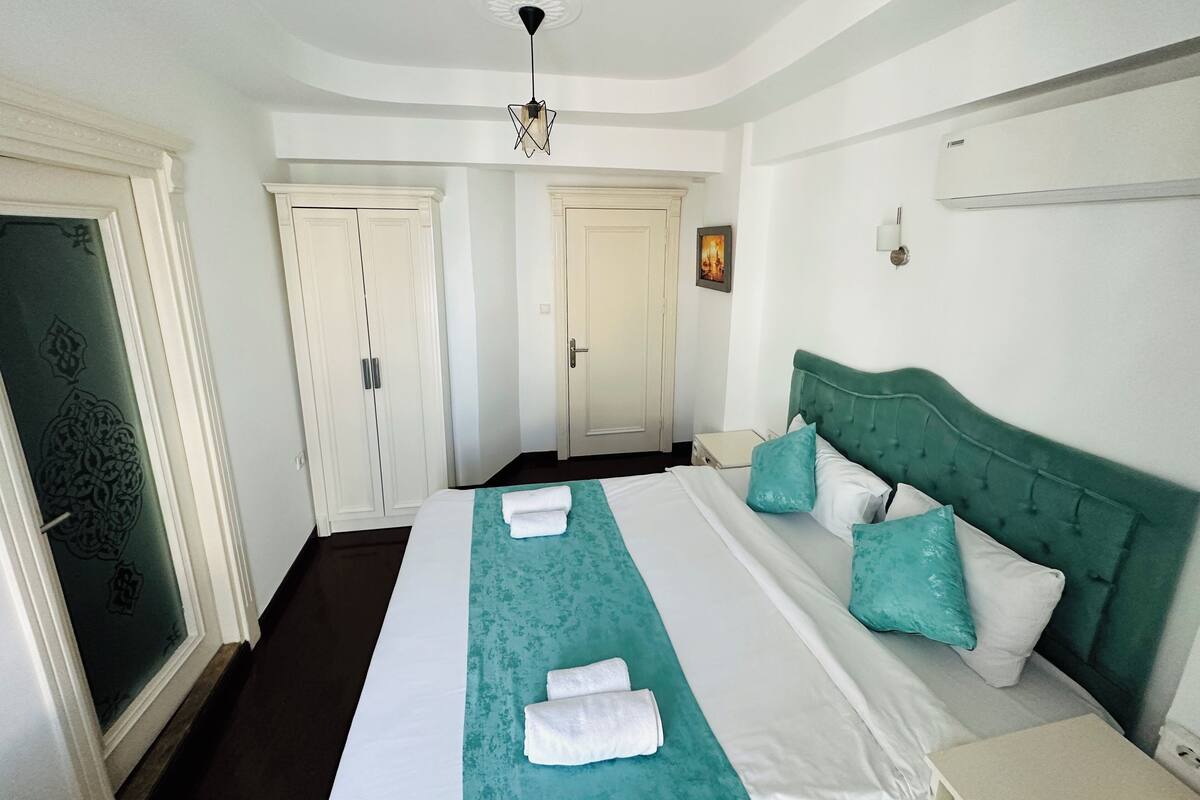 A hotel room with a large bed, two white doors, a green headboard, and a ceiling light fixture.