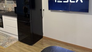 A modern kitchen with a black refrigerator, a tiled backsplash, and a wall-mounted TV displaying the Tesla logo. - Le Mini Versailles (Goussainville)