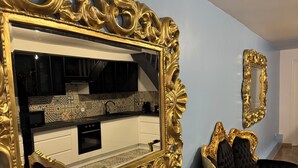 A room with a large ornate gold-framed mirror, a black upholstered chair, and a kitchen visible in the reflection. - Le Mini Versailles (Goussainville)