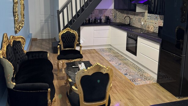 A modern living room with a staircase, a kitchen with a black refrigerator, and a dining area with black and gold chairs. - Le Mini Versailles (Goussainville)