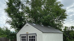 A small, single-story shed with a gray door and windows, surrounded by a wooden fence and greenery.