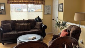 A living room with a sofa, a coffee table, and a chair. There is a window with a view of a house and a door.