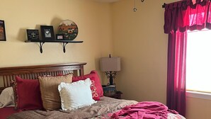 A bedroom with a wooden bed, a ceiling fan, a nightstand with a lamp, a shelf with framed items, and a window with red curtains.