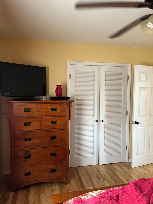 Une chambre avec une commode en bois, un téléviseur, un vase rouge et des portes à persiennes blanches.