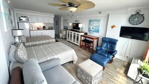 A modern hotel room with a bed, a blue armchair, a small table, and a ceiling fan. - Spacious Oceanfront Studio w/2 queen beds & sleeper, kitchenette, pool, tiki bar (Daytona Beach)