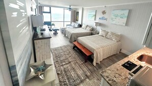 A modern hotel room with two beds, a kitchenette, a large window with a view of the sea, and a ceiling fan. - Spacious Oceanfront Studio w/2 queen beds & sleeper, kitchenette, pool, tiki bar (Daytona Beach)