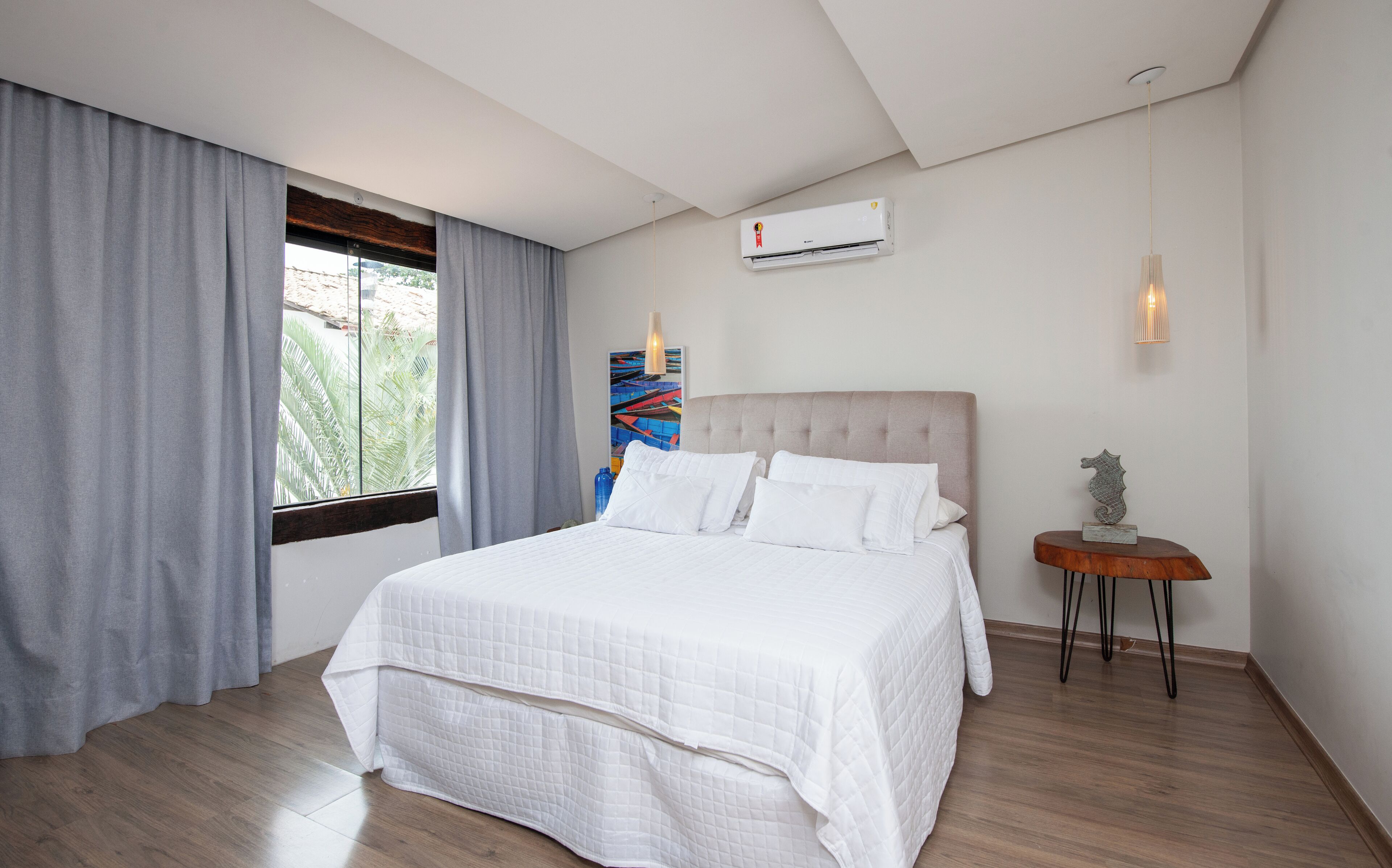 Une chambre d’hôtel moderne avec un grand lit, des rideaux gris, une fenêtre donnant sur un beau paysage, un appareil de climatisation et une petite table agrémentée d’un objet décoratif.