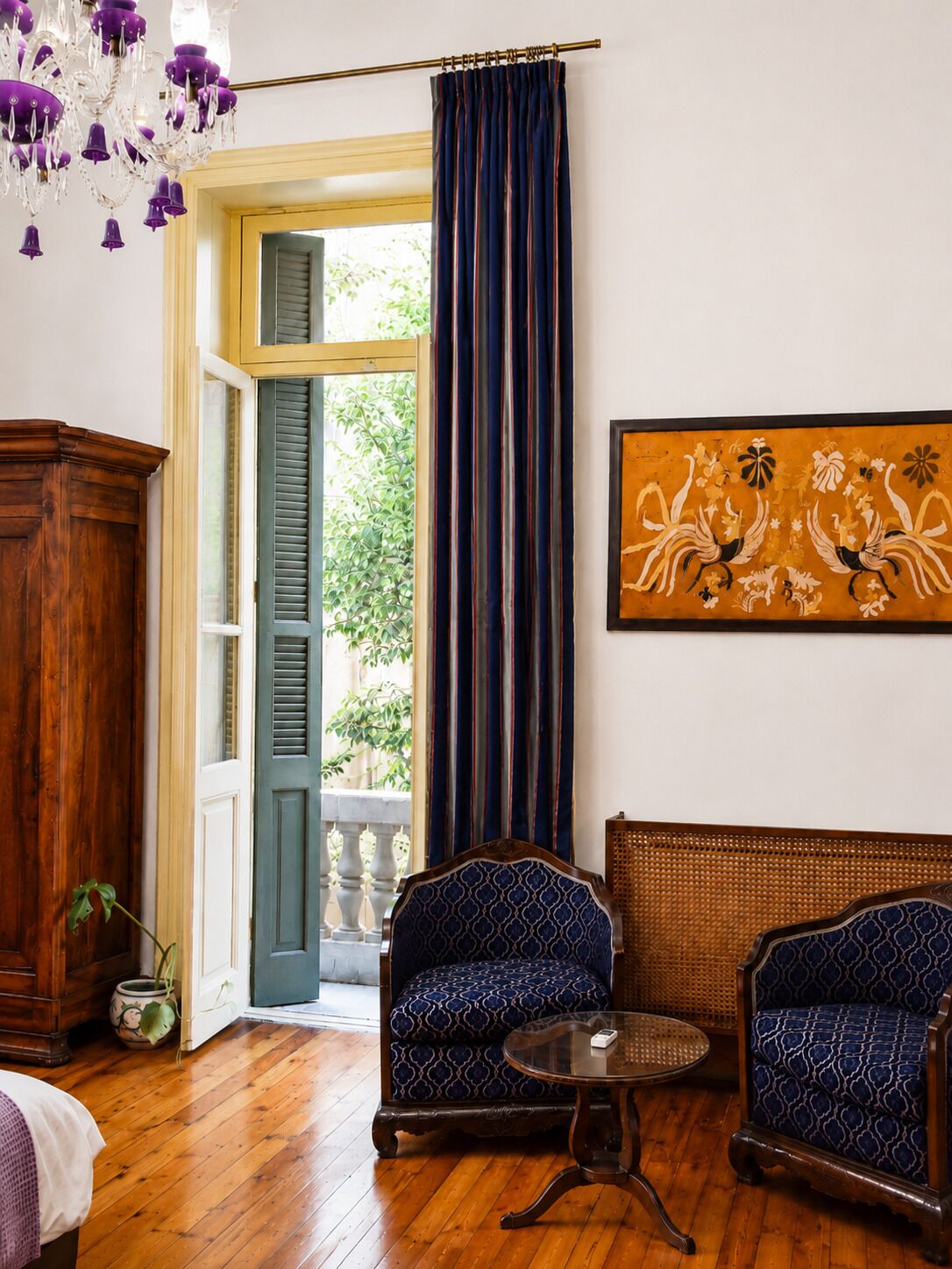 Quarto com piso de madeira, duas poltronas estampadas, uma pequena mesa redonda, uma pintura grande e vista através de uma porta com persianas.