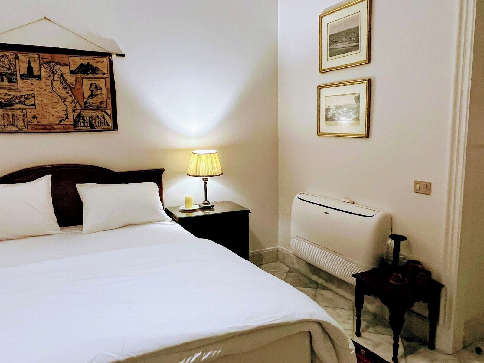 Een hotelkamer met een bed, een nachtkastje met een lamp, een wandairconditioner en ingelijste foto's.
