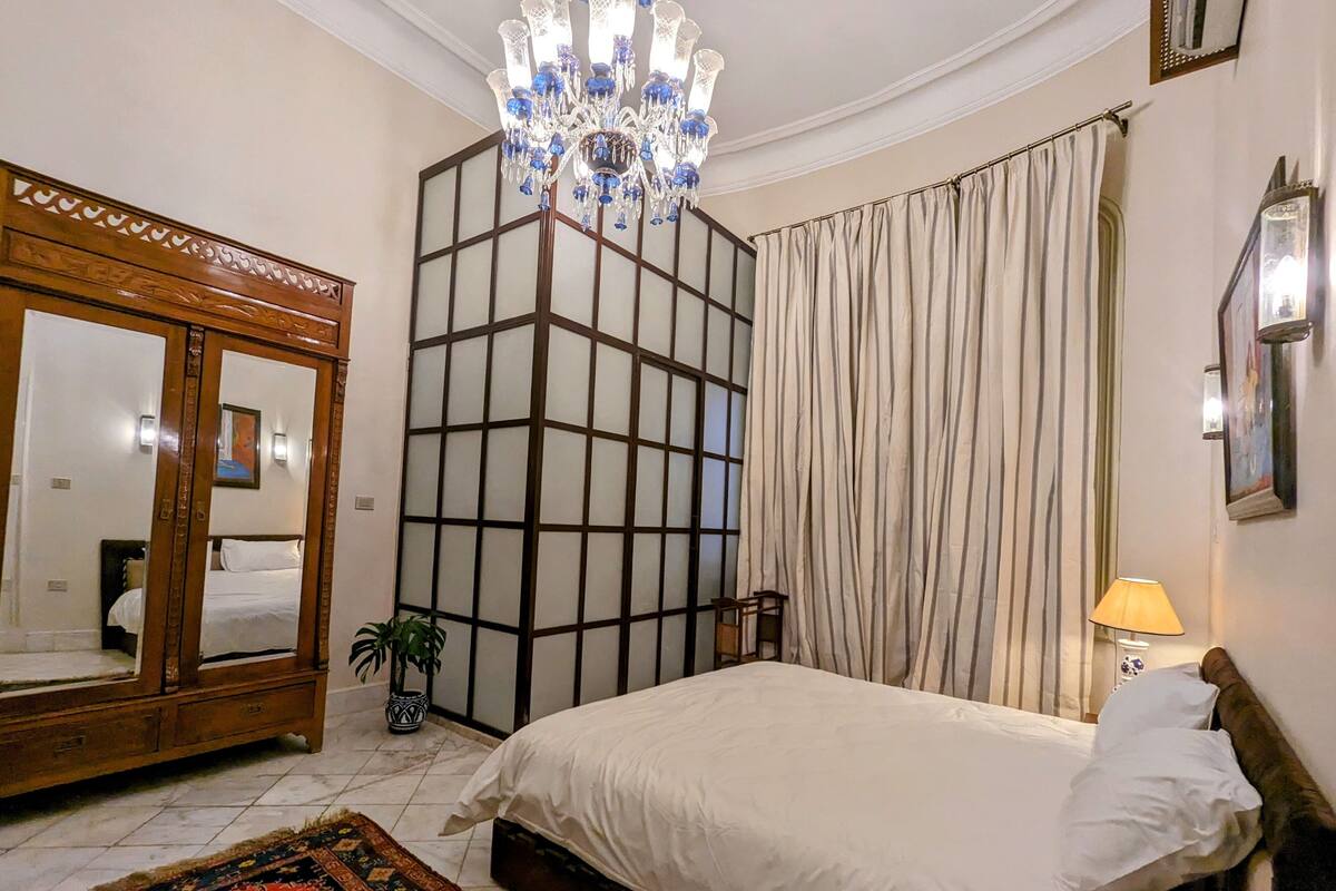 A bedroom with a large bed, a wooden wardrobe, a mirror, a chandelier, a potted plant, and a window with curtains.